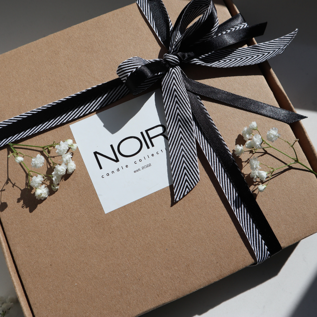 Gift box with a black and white ribbon and 'NOIR' label, surrounded by small white flowers.